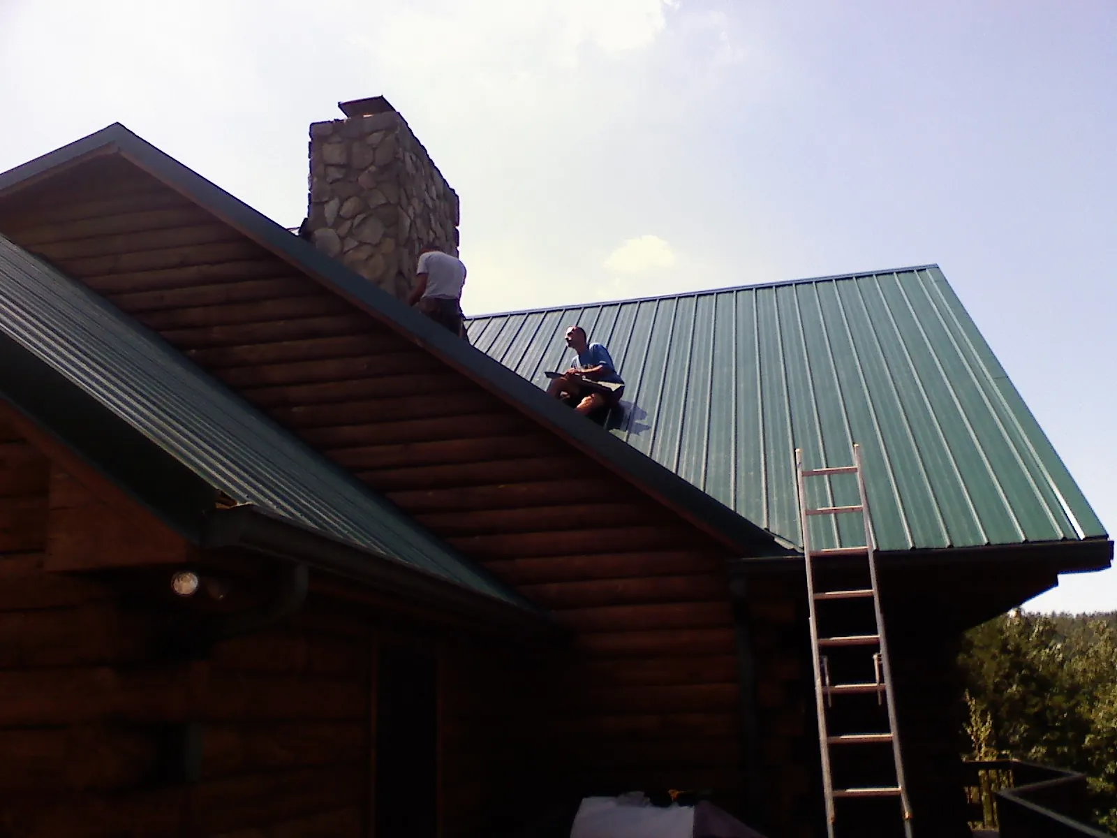 Roofers installing green metal panels beside a stone chimney on a log home in San Clemente